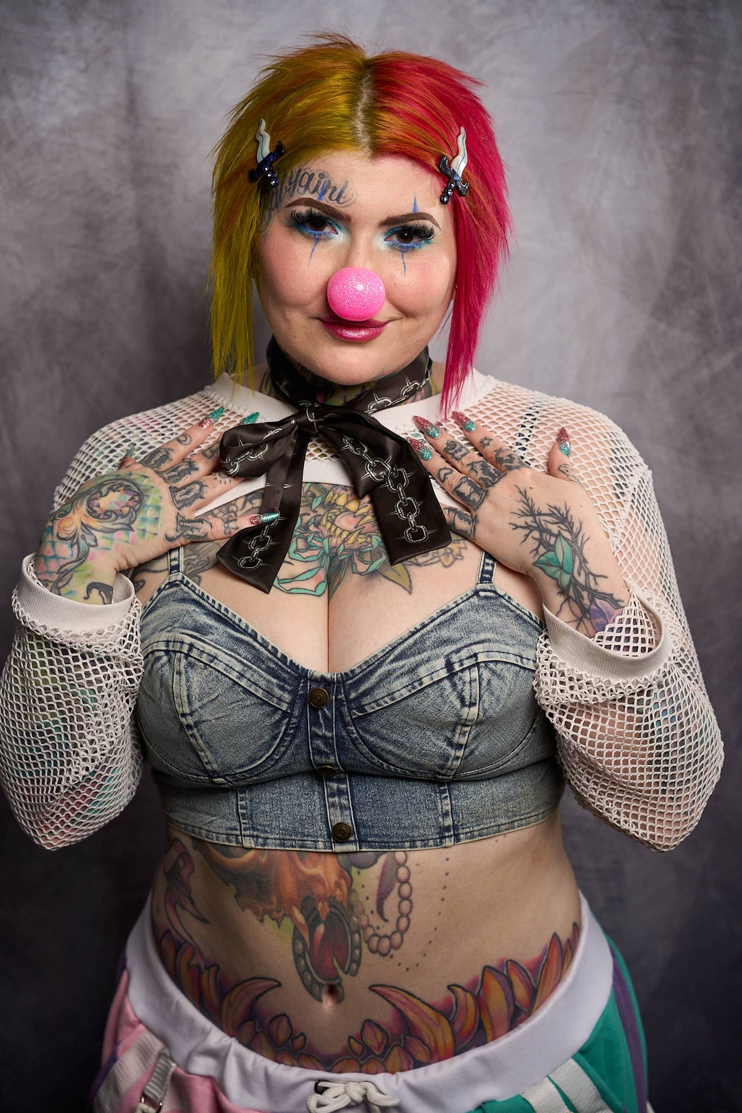 Person with colorful hair and tattoos wearing a denim outfit against a gray background