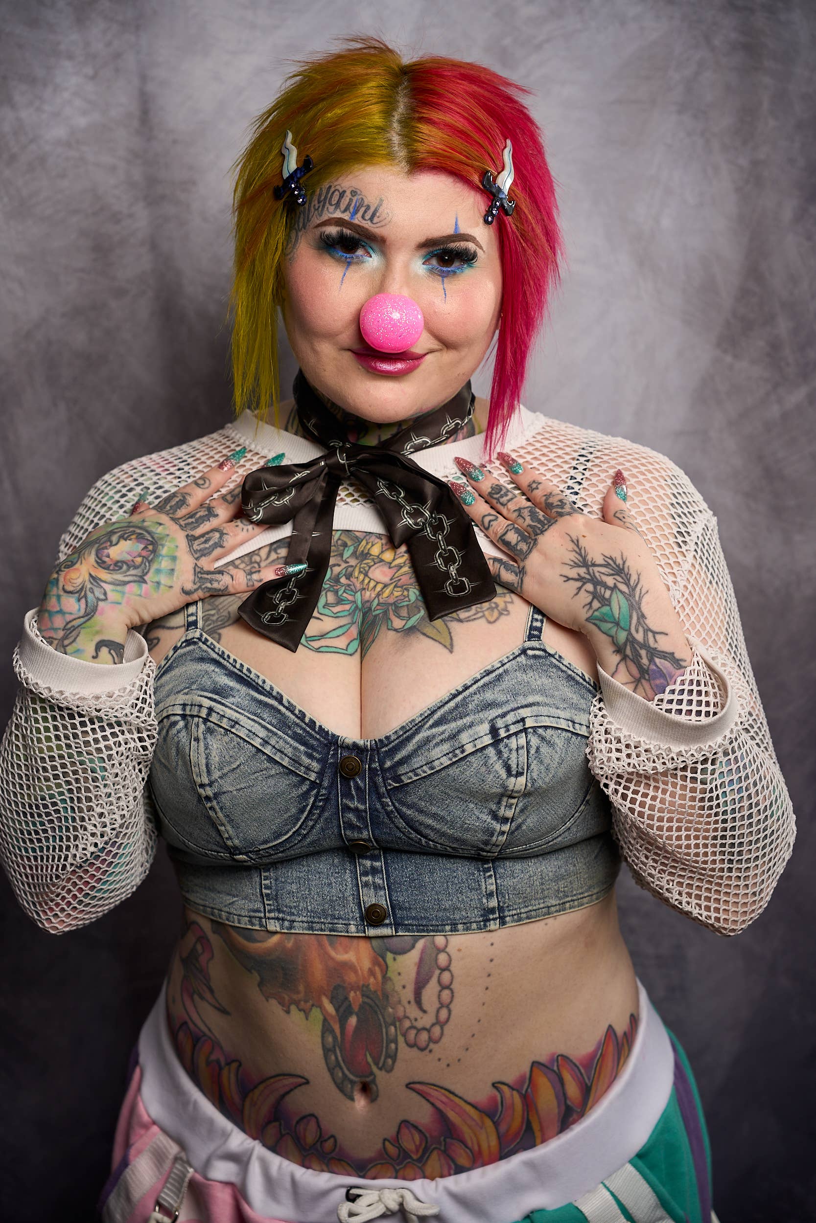 Person with colorful hair and tattoos wearing a denim outfit against a gray background