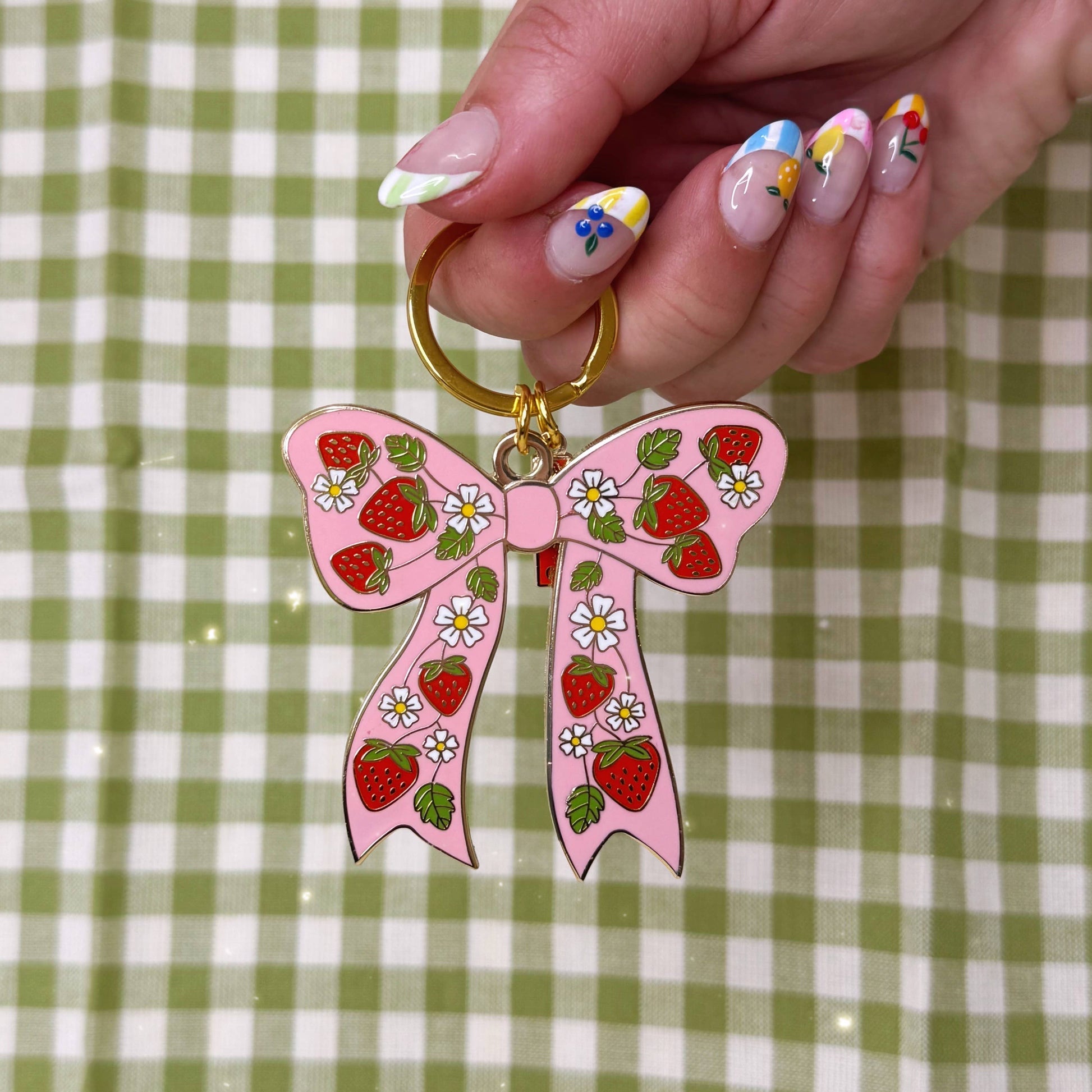 Pink bow keychain with strawberry design held by a hand on a green and white checkered background