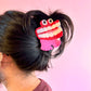 Hair clip shaped like a cartoonish toothy monster on a person's hair against a pink background