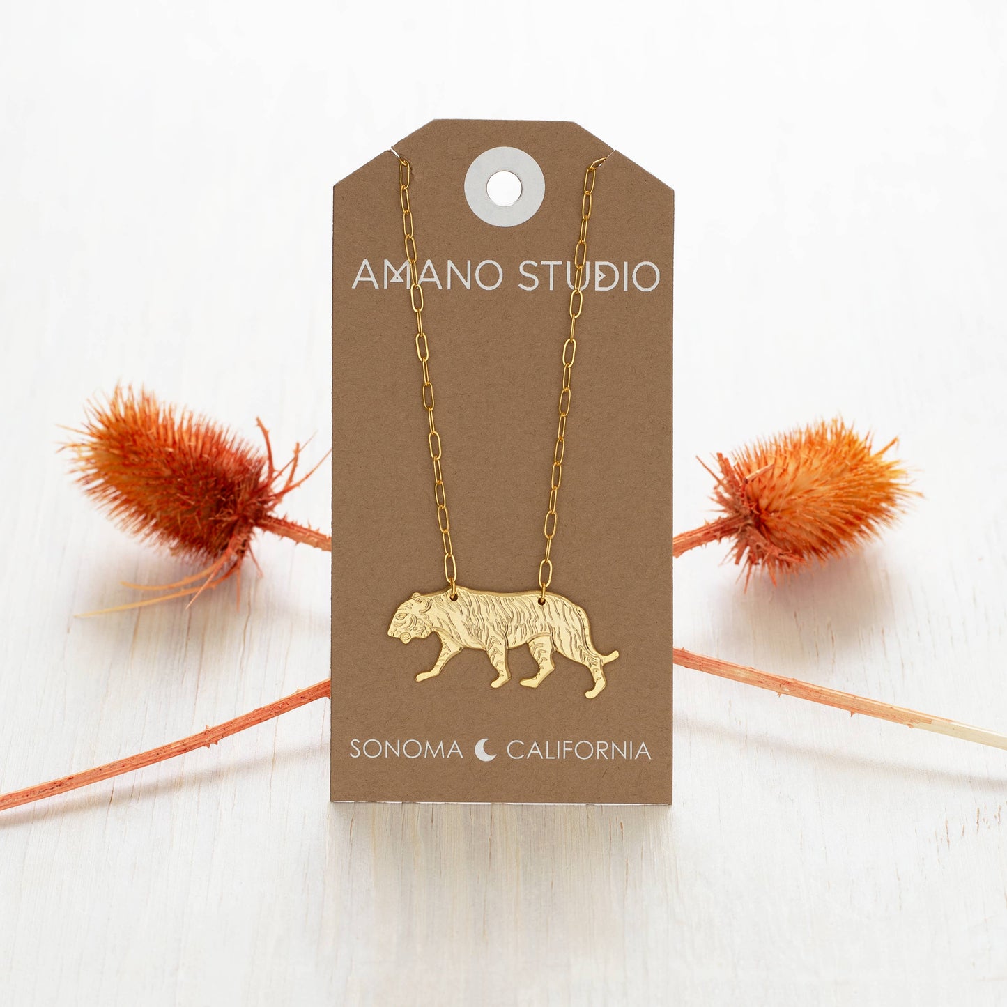 Gold necklace with a tiger pendant on an Amano Studio card with decorative elements.