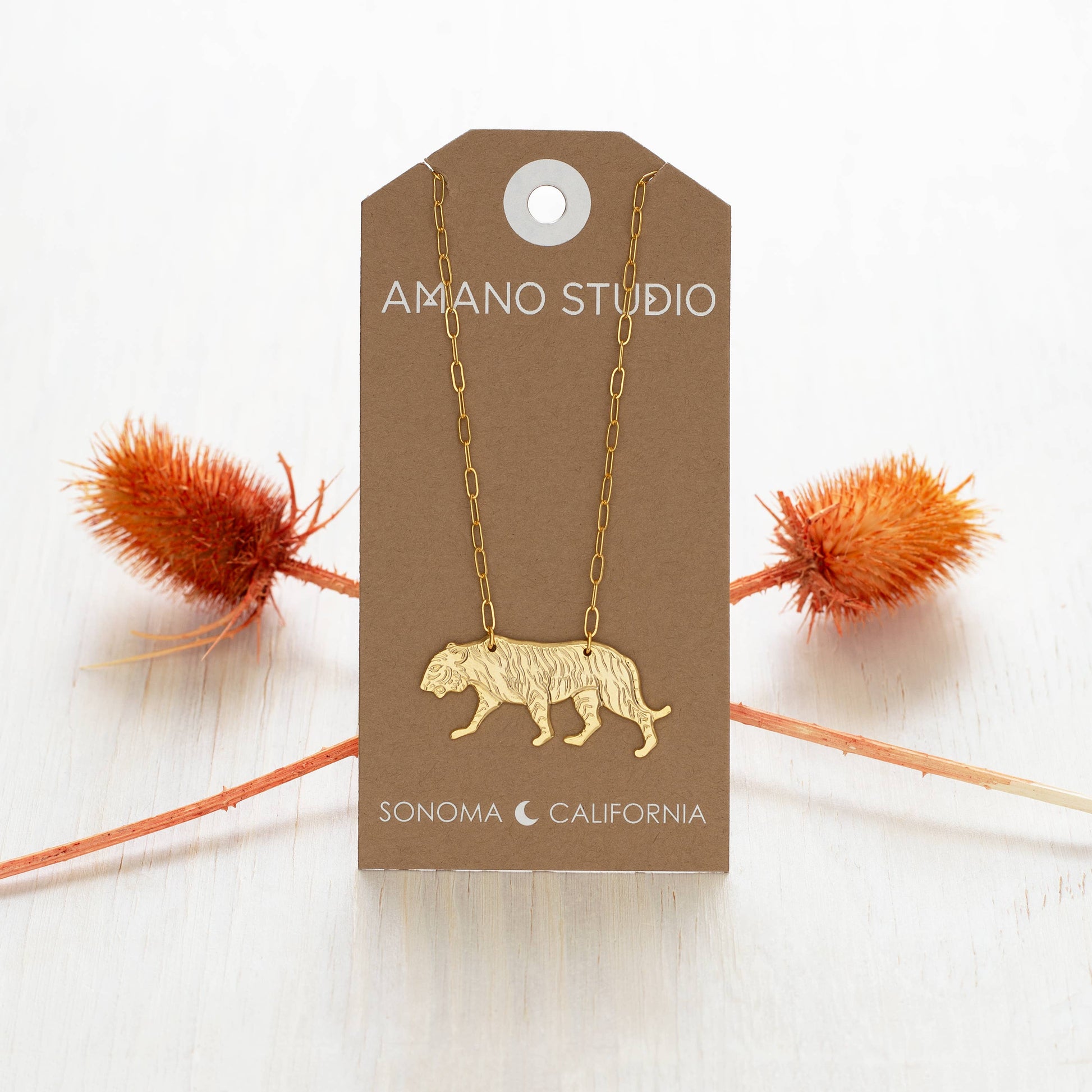 Gold necklace with a tiger pendant on an Amano Studio card with decorative elements.