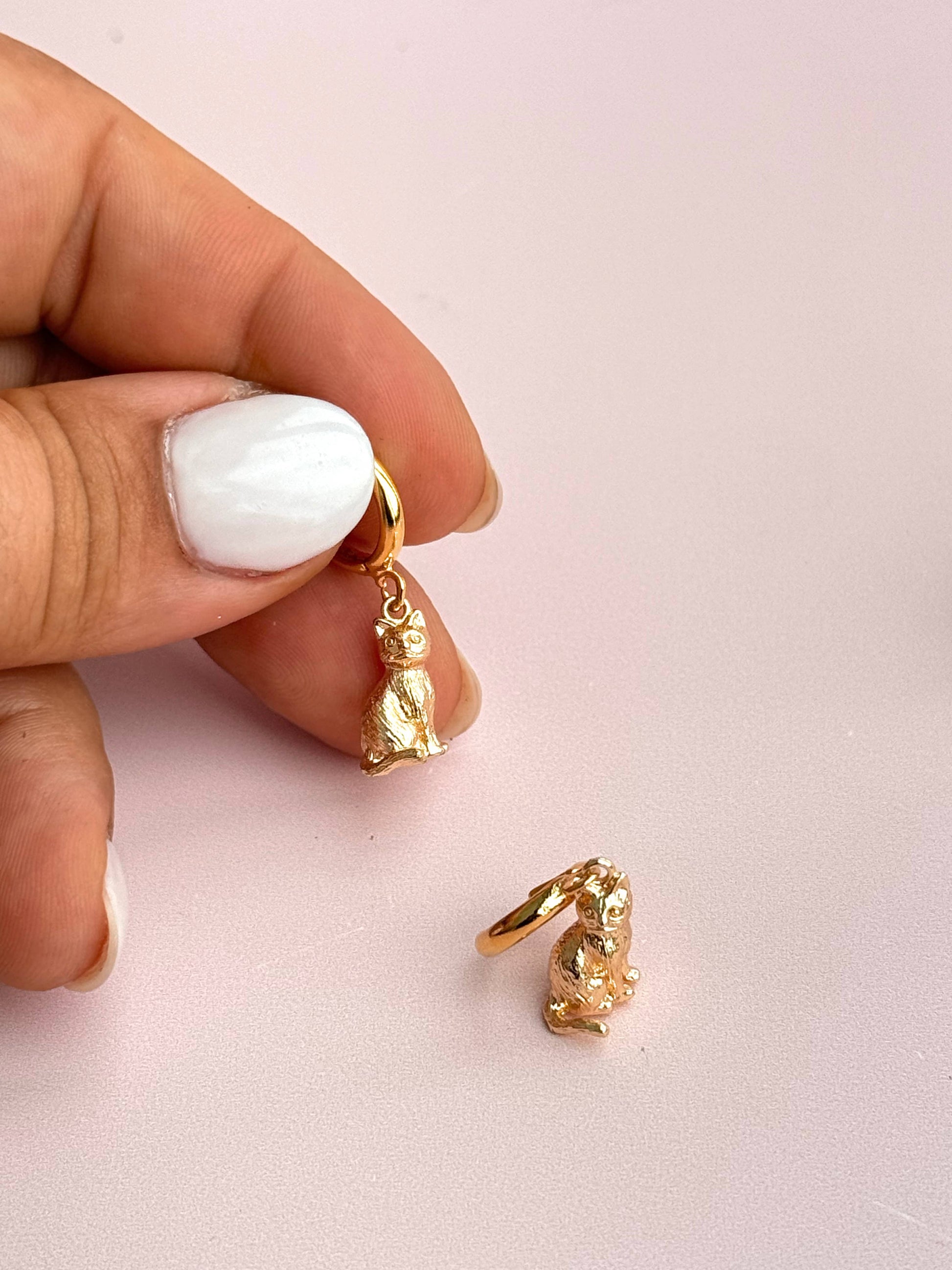 Gold cat hoop earrings held between fingers against a light pink background