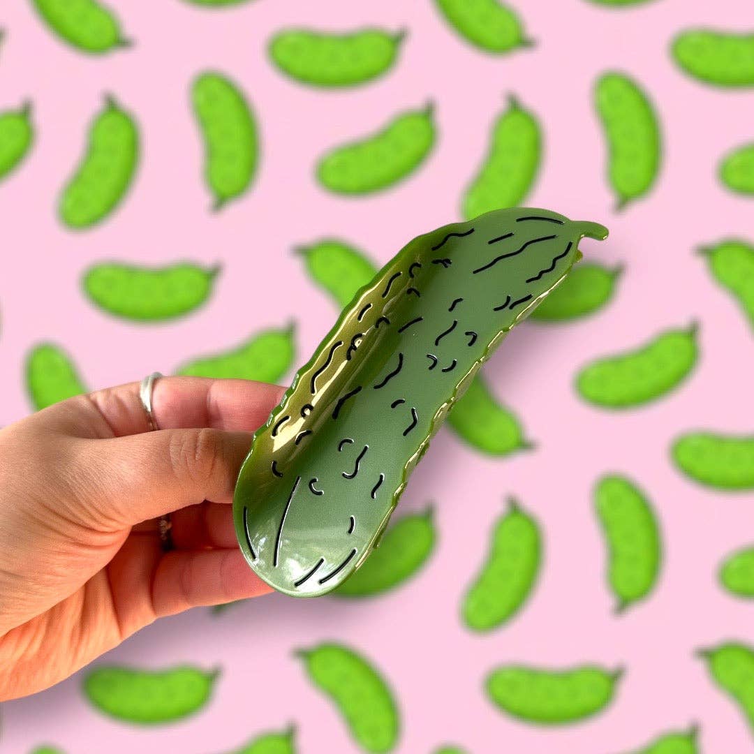 Hand holding a green pickle-shaped hair claw against a pink background with green pickle patterns.