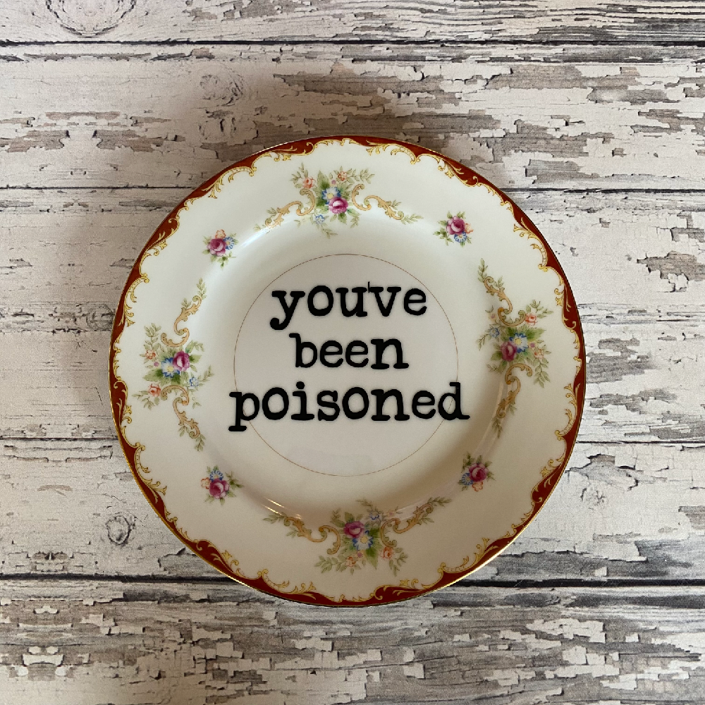 Decorative plate with floral patterns and text 'you've been poisoned' on a wooden surface.