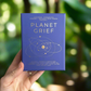 Hand holding a blue book titled 'Planet Grief' against a blurred natural background